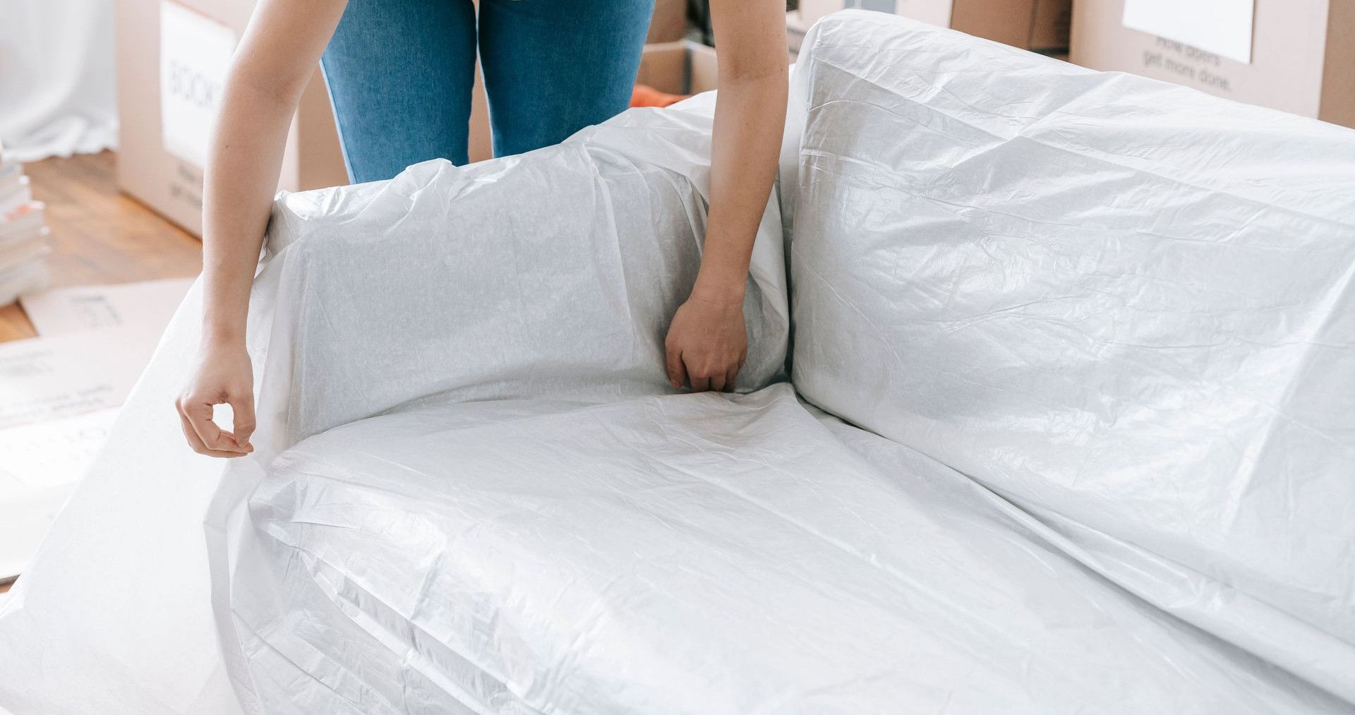 A woman carefully wraps a sofa in protective material while packing boxes in a bright room.